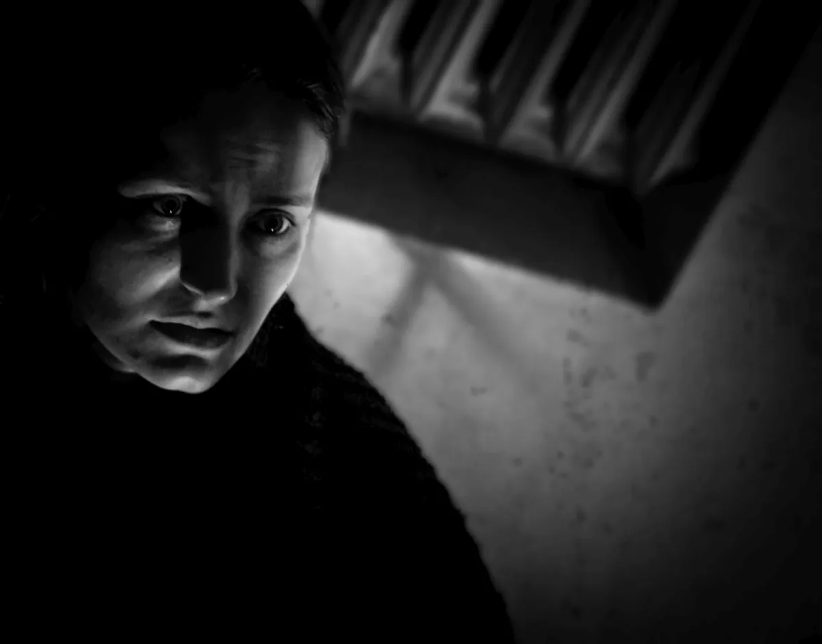 Young woman lit from one side in deep shadow beneath a shelf of books